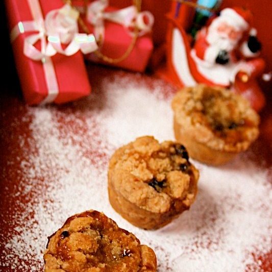 Mini Almond Crumble Mince Pies: on a red background with dusting of icing sugar like snow. In soft focus are dinky presents and a santa ornament.