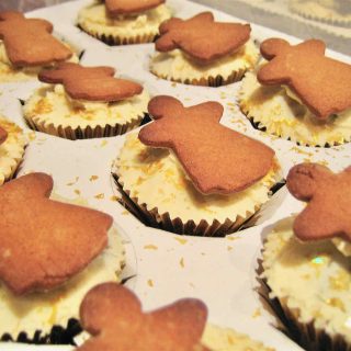 Vanilla buttercream topped cupcakes in a cardboard tray decorated with angel shaped gingerbread biscuits and scattered with gold edible dust