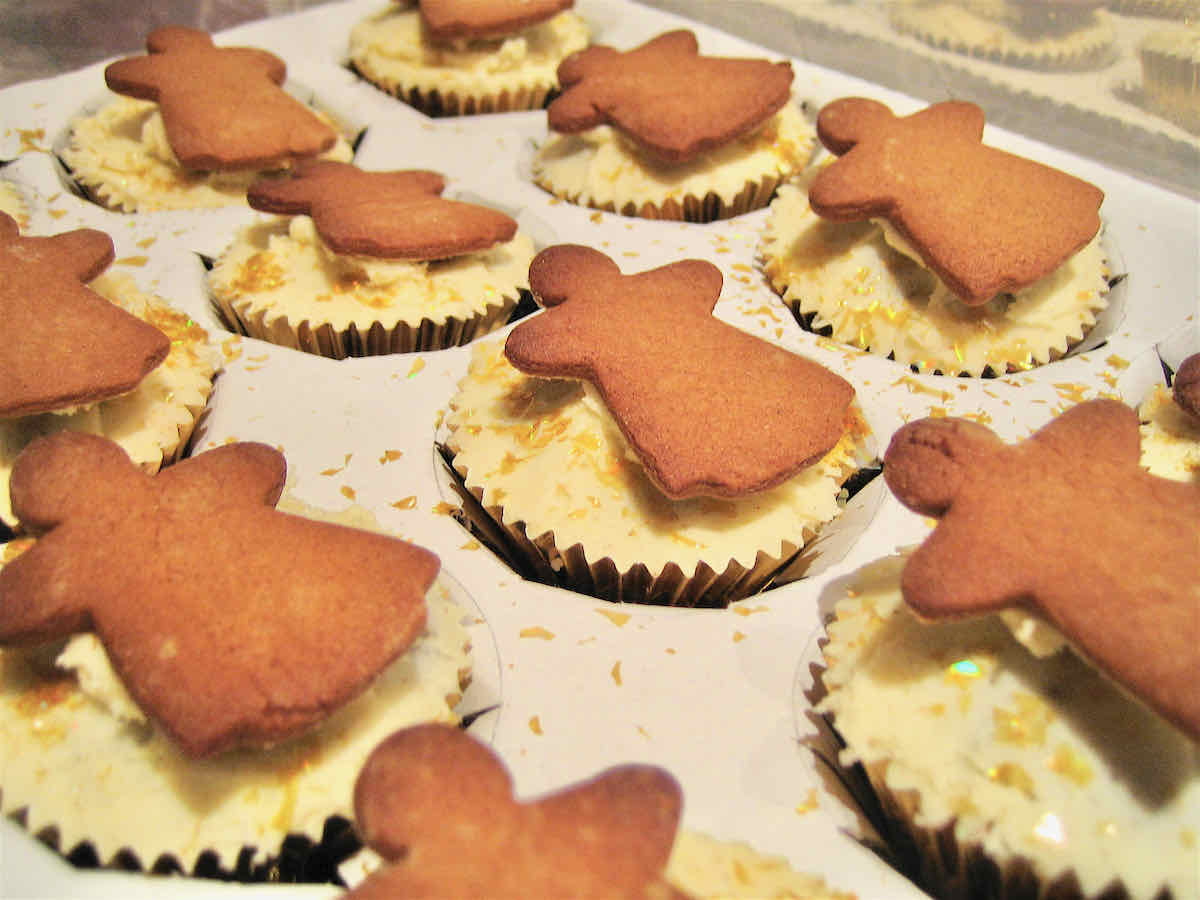 Vanilla buttercream topped cupcakes in a cardboard tray decorated with angel shaped gingerbread biscuits and scattered with gold edible dust