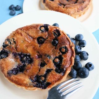 A golden blueberry tart on a white plate with raw blueberries and a fork.