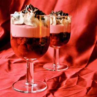 Two stem glasses filled with raspberry trifle in layers of red jelly, pink custard, cream and chocolate sprinkles.