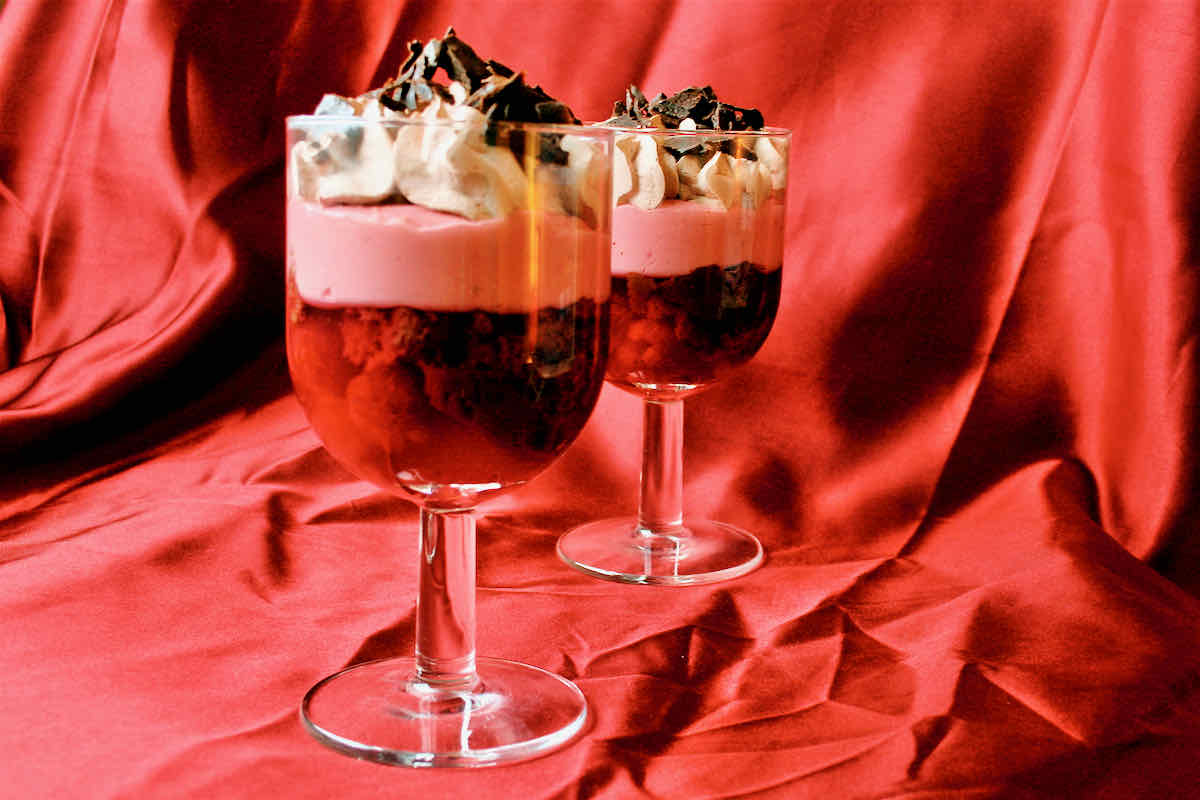 Two stem glasses filled with raspberry trifle in layers of red jelly, pink custard, cream and chocolate sprinkles.