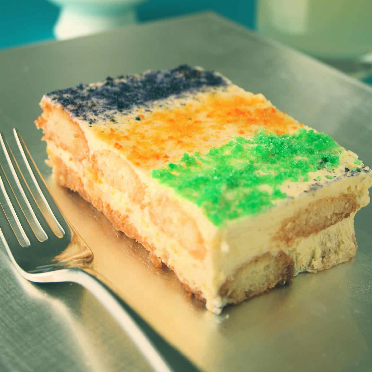 A rectangular slice of tiramisu covered with purple, orange and green sugars in the manner of New Orleans King Cake