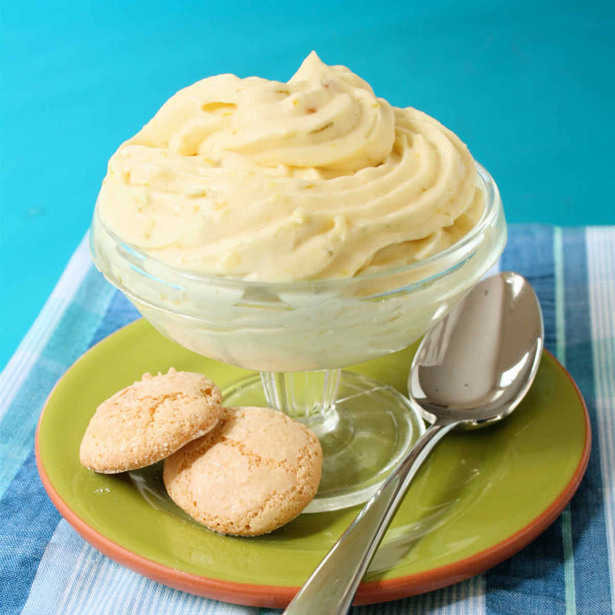 A stemmed dessert glass filled with piped pale yellow mousse on a green plate with spoon and two small round biscuits.