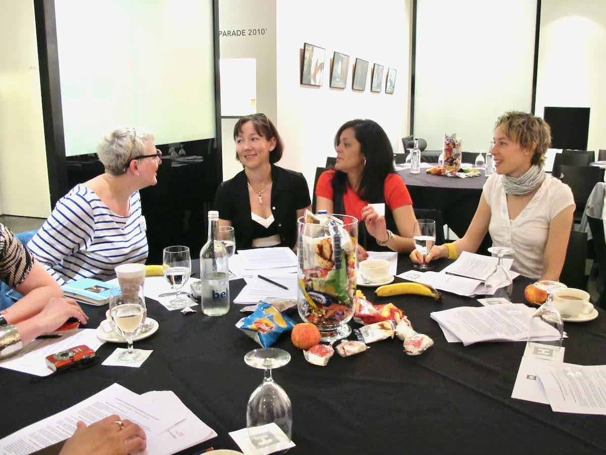 Four ladies at a conference table in conversation, there are snacks and drinks on the table and a black tablecloth. The background is white and brightly lit. 