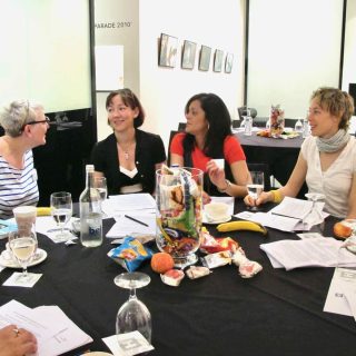 Four ladies at a conference table in conversation, there are snacks and drinks on the table and a black tablecloth. The background is white and brightly lit.