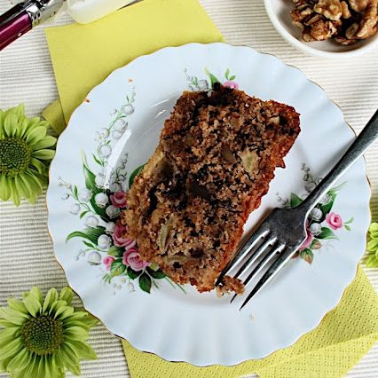 Apple Loaf Cake with Nuts and Chocolate: from above, a white plate with dessert fork, rectangle slice of loaf cake, on a surface with green flowers and paper napkin.