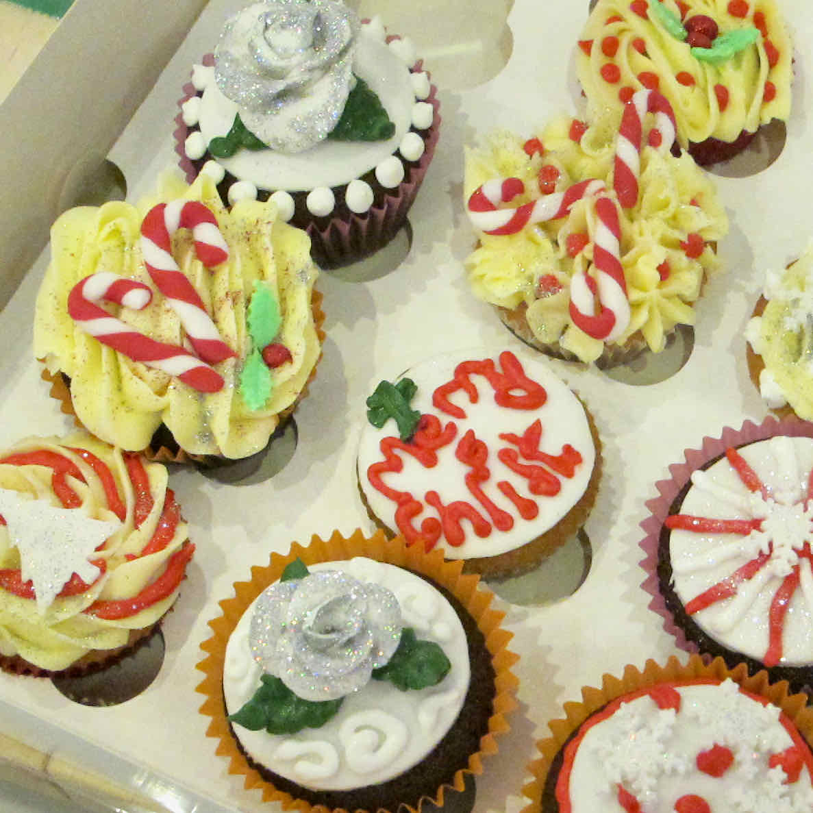 Mich Turner Cupcake Class: Selection of hand decorated Christmas themed cupcakes with glittery and messy decoration in a cardboard box.