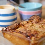 Golden bread and butter pudding in glass dish and in soft focus background are shown a blue and white striped jug and some stacked pastel pink and blue bowls.