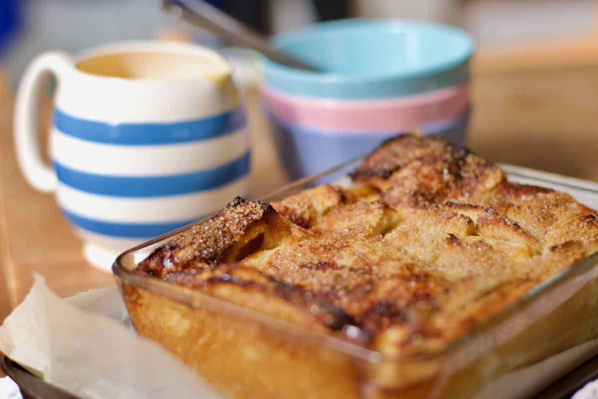 Golden bread and butter pudding in glass dish and in soft focus background are shown a blue and white striped jug and some stacked pastel pink and blue bowls.