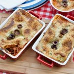 Two individual sized Black Olive Lasagnes baked in individual red and white oven dishes upon a wooden board on a red white gingham table cloth