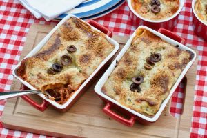 Two individual sized Black Olive Lasagnes baked in individual red and white oven dishes upon a wooden board on a red white gingham table cloth