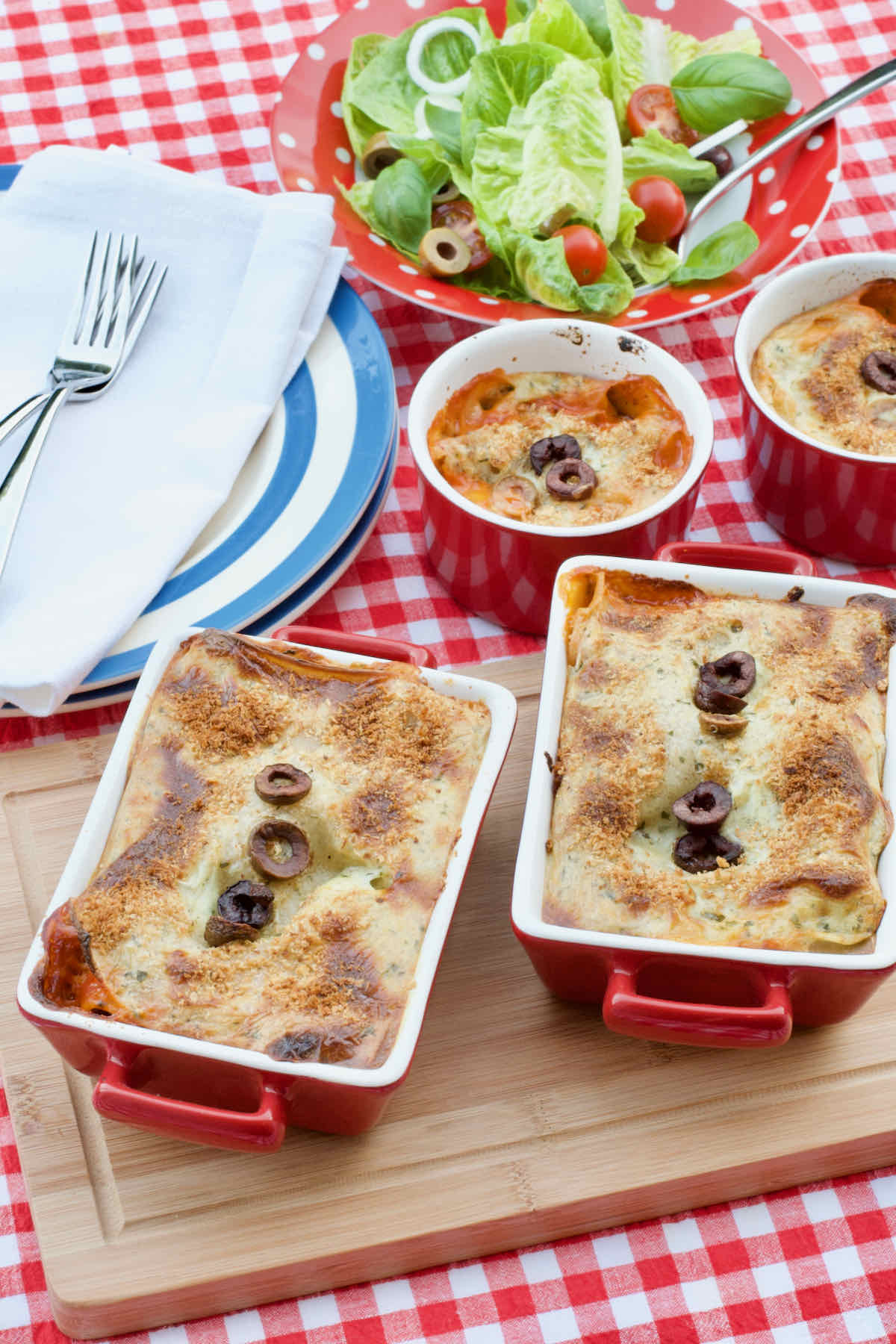 Two individual sized Black Olive Lasagnes baked in individual red and white oven dishes upon a wooden board on a red white gingham table cloth, a blue plate and bowl of salad lie behind