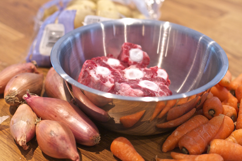 Oxtail Beef Bones used in Crockpot Beef on the Bone Stew: beef bones in a stainless steel bowl surrounded by shallots and carrots.