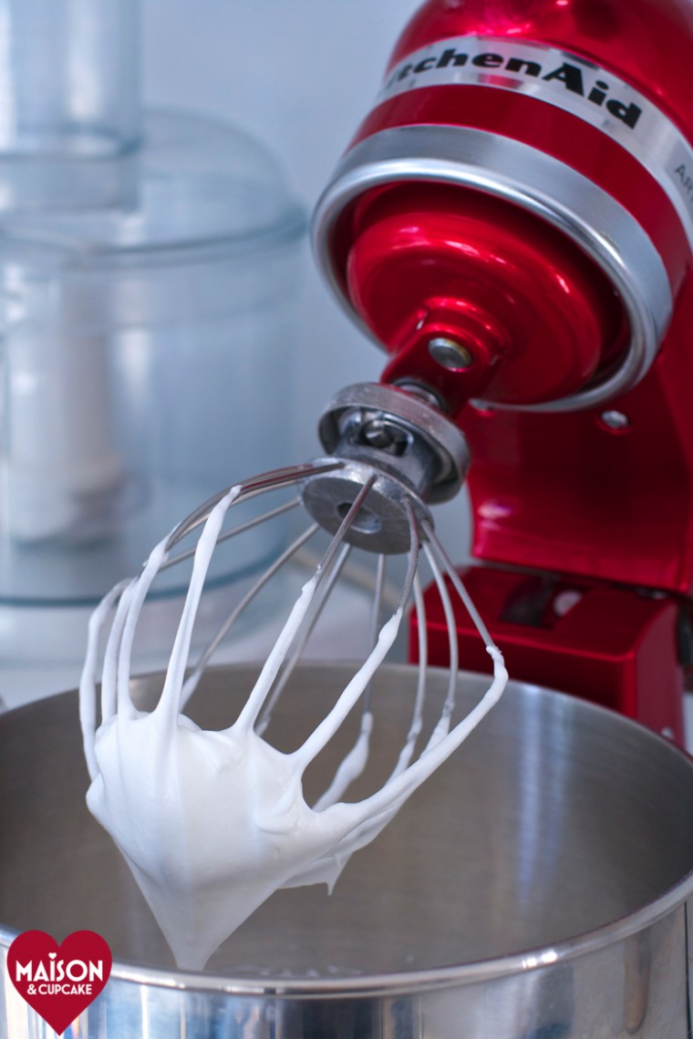 Whisk attachment on red KitchenAid food mixer; whisked egg whites are on the lifted blades above stainless steel bowl