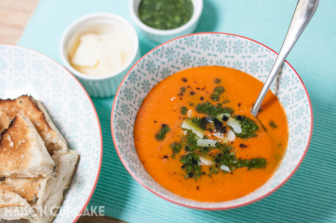 Roast tomato and red pepper soup topped with basil oil and shaved grana padano cheese served in pattern light blue bowls, smaller white dishes of the toppings are to the side. Toasted bread is on another plate to the left.