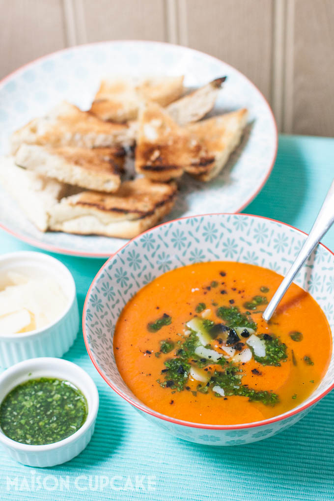 Roast tomato and red pepper soup topped with basil oil and shaved grana padano cheese served in pattern light blue bowls, smaller white dishes of the toppings are to the side. In soft focus, toasted bread is on another plate behind.