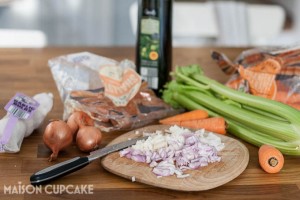 A small black knife on a wooden chopping board with finely chopped shallots; whole shallots, celery sticks and small carrots lie around this and plastic vegetable packaging and an olive oil bottle are in the background.