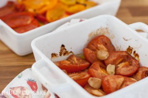 Two white square oven dishes filled with roast tomatoes, garlic cloves and red and orange pepper strips.