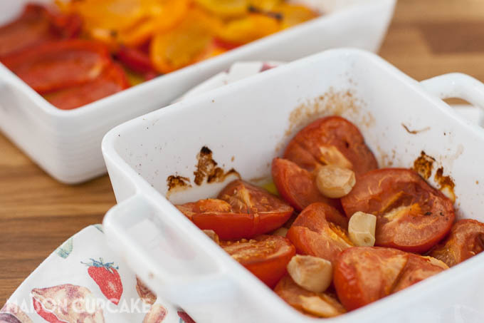 Two white square oven dishes filled with roast tomatoes, garlic cloves and red and orange pepper strips.