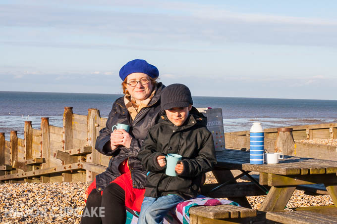 Whitstable's pebbled beach and groynes on a bright winter's day; a lady in a blue beret and a young boy in black peaked cap hold light blue plastic mugs of soup. A striped vintage flask sits on the wooden picnic table beside them. 