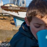 On Whitstable pebble beach on a bright winters day, a small boy sips soup from a pale blue mug, his face half covered in shadow.