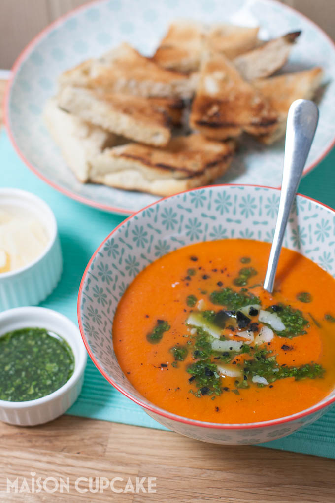 Roast tomato and red pepper soup topped with basil oil and shaved grana padano cheese served in pattern light blue bowls, smaller white dishes of the toppings are to the side. In soft focus, toasted bread is on another plate behind.