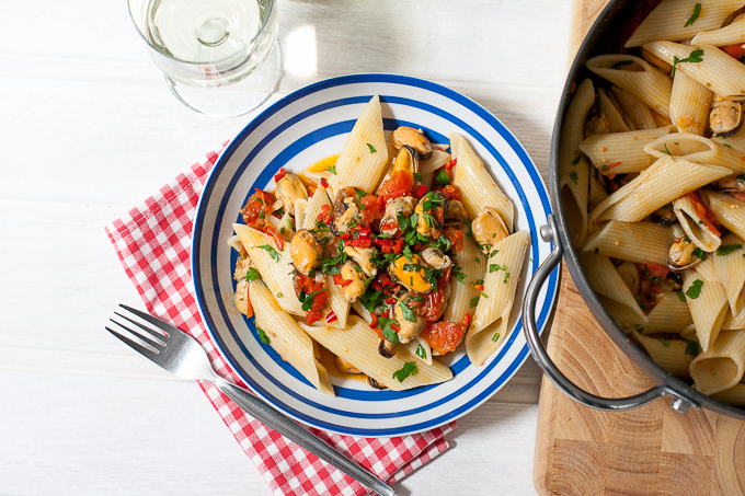 Pennoni with Chilli Mussels giant pasta recipe: in a blue striped bowl is served giant penne pasta quills with mussels and chopped herbs and tomatoes. A fork and check red white napkin are underneath.