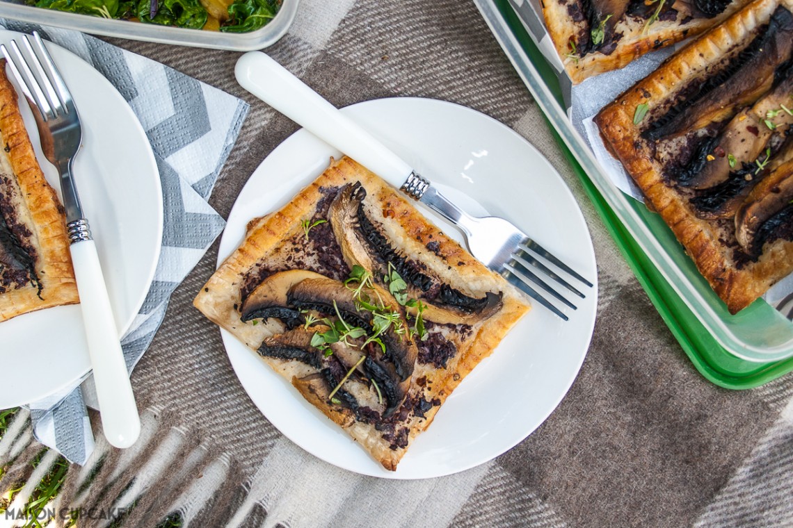 Portobello mushroom tarts recipe with black olive tapenade. From above, a square mushroom tart slice on a white plate upon a grey check picnic blanket. Picnicware and other plates are partly in shot.