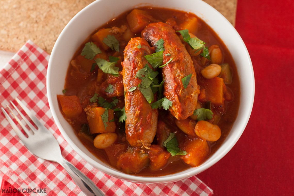 A white bowl of low fat sausage casserole with beans in tomato sauce upon a red gingham napkin with a fork.