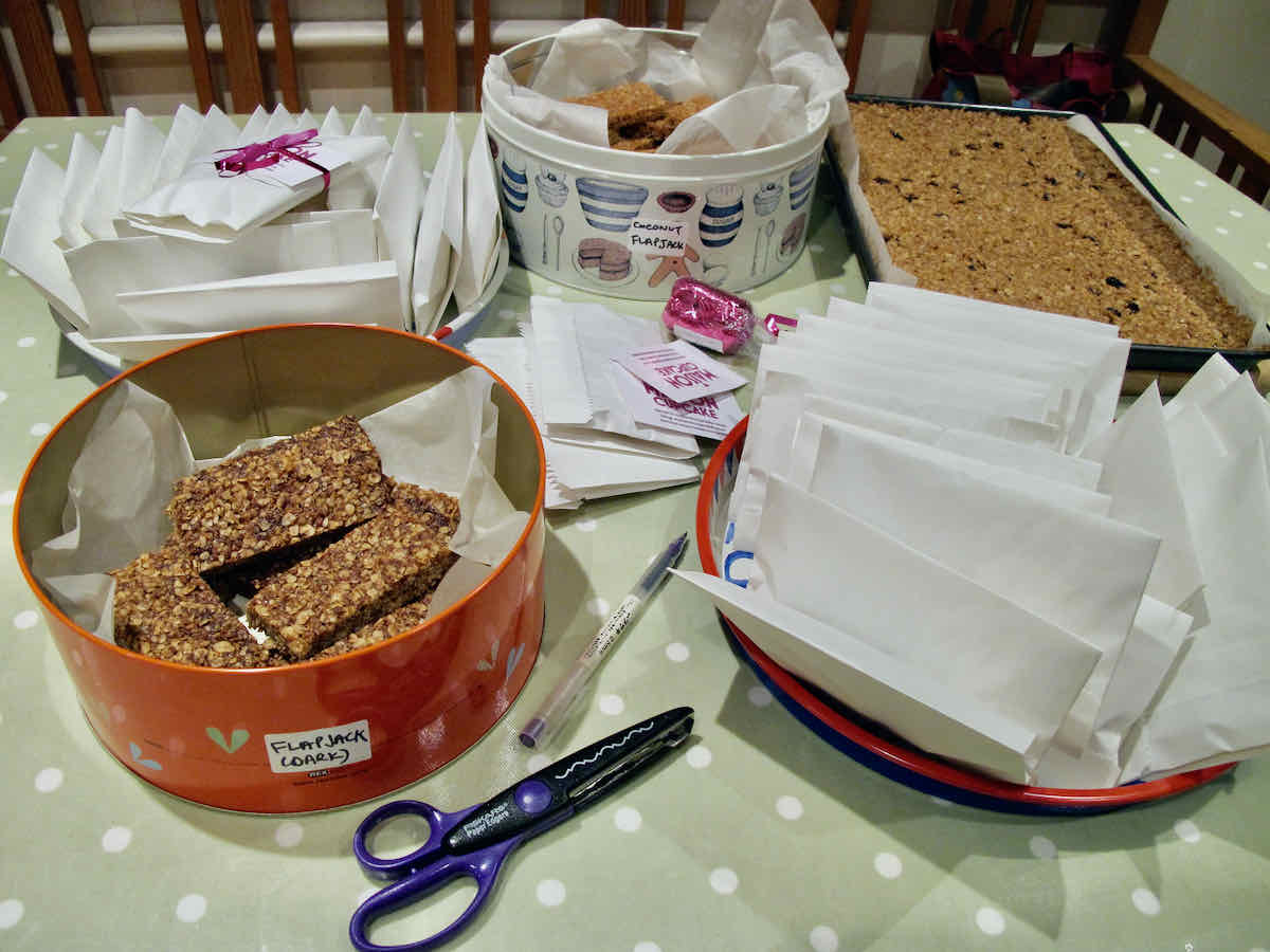 A tabletop scene wrapping flapjacks into parcels; with scissors, ribbon, a tin of flapjacks and white paper bags.