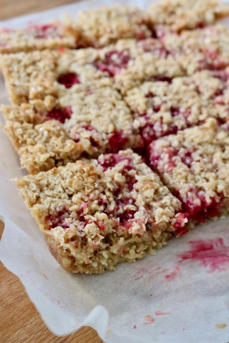 Raspberry and Coconut Flapjacks cut into squares on baking paper. Red raspberry fruit is visible and has stained the paper.