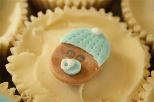 Baby shower cupcake decorated with cream buttercream icing and a sugarpaste baby face with bobble hat and dummy.
