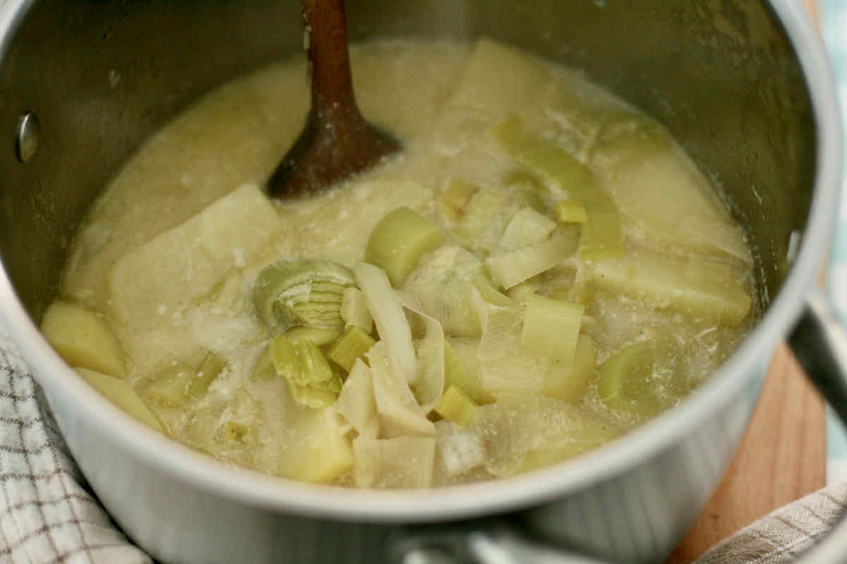 Steps making easy leek and potato soup - the pale yellow soup in a steel pan with wooden spoon before blending. Cooked leeks are visible.