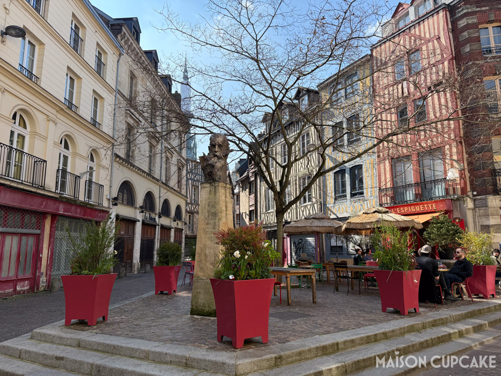 Place Saint-Amand in Rouen with red planters, bust of Claude Monet and L'Espiguette cafe umbrellas and tables.